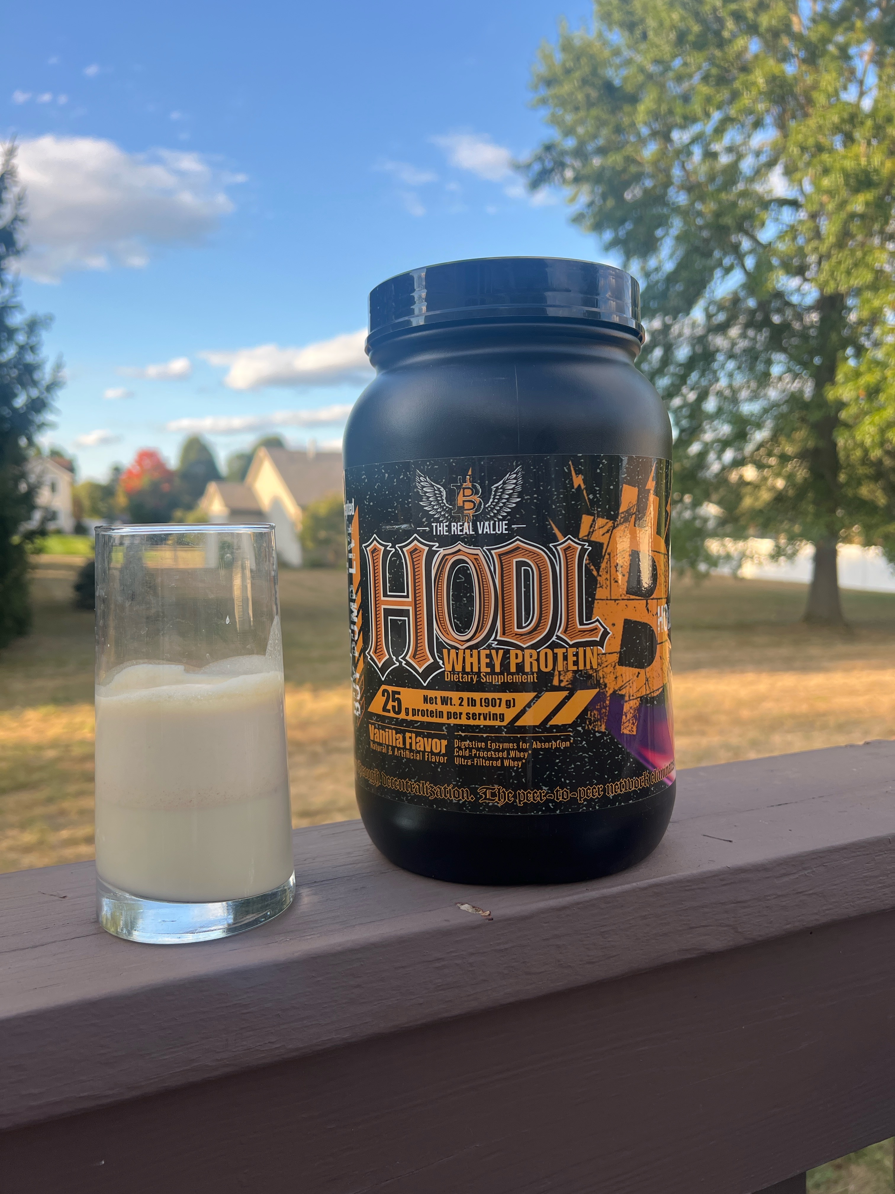 A bottle of HODL THE WHEY protein and a glass of raw milk with peanut butter placed on an outdoor porch railing.